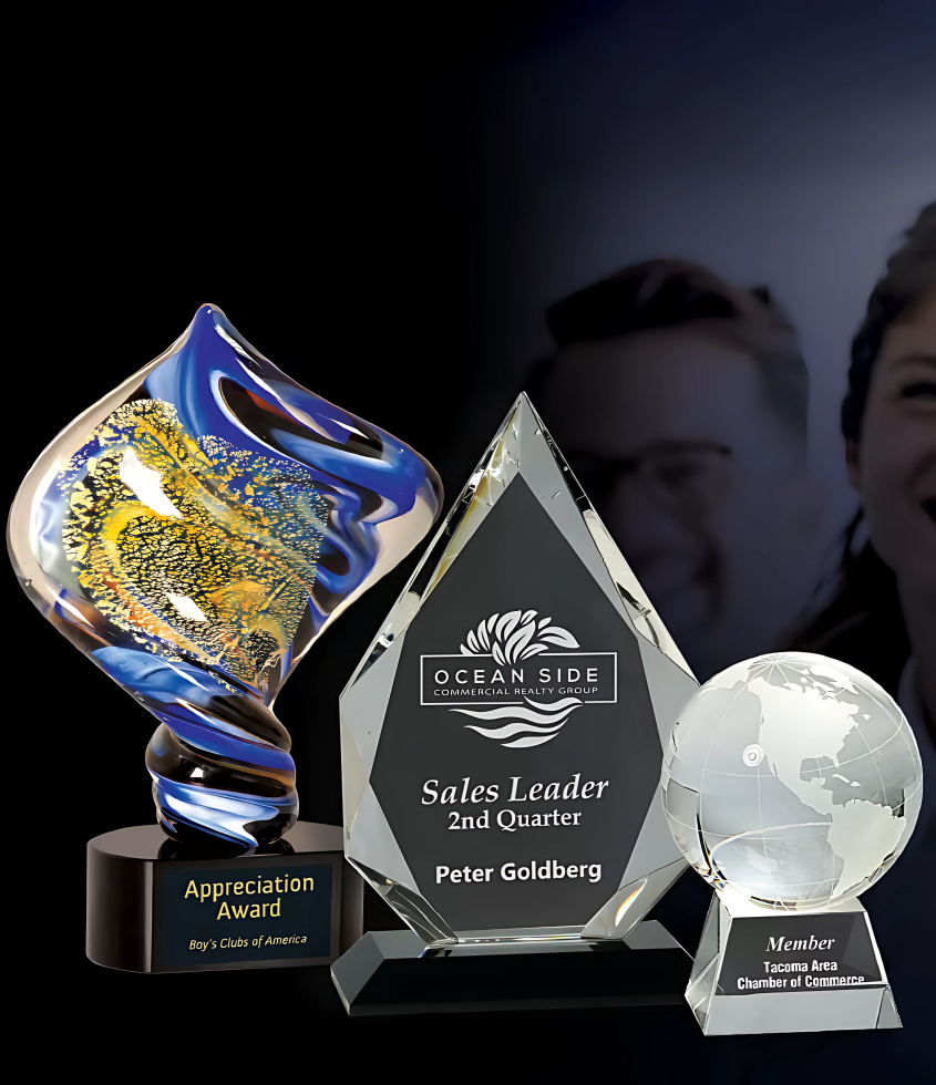 Display of three corporate awards: a decorative blue and gold art glass trophy, a crystal diamond-shaped sales achievement plaque, and a crystal globe award.